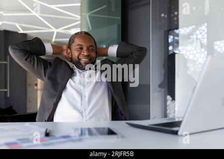 Ein gut gefertigter Geschäftsmann hat die Arbeit erfolgreich abgeschlossen, ruht träumerisch und nachdenklich im Büro, der Mann wirft seine Hände hinter den Kopf und visualisiert die zukünftigen Siege und Erfolge. Stockfoto