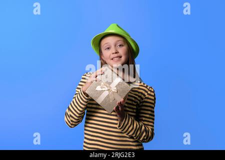 Ein Mädchen mit einer Geschenkbox in den Händen und einem grünen Karnevalsmütze lächelt und schaut auf die Kamera. Isoliert auf hellblauem Hintergrund Stockfoto