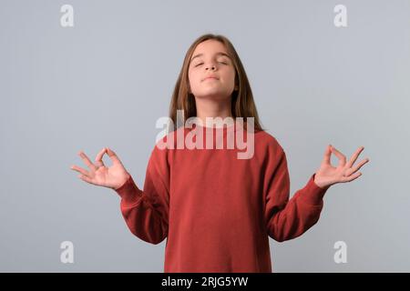 Schönes Teenager-Mädchen, das roten Pullover auf blauem Hintergrund trägt, Yoga macht, die Augen geschlossen hält, die Finger in Mudra-Geste hält. Meditation Stockfoto