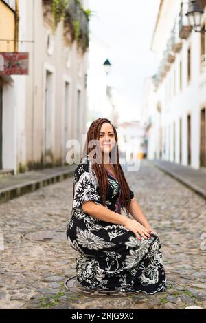 Eine Frau hockte auf einer Kopfsteinpflasterstraße. Haargeflechte und afrikanisches Kleid. Pelourinho, Brasilien. Stockfoto