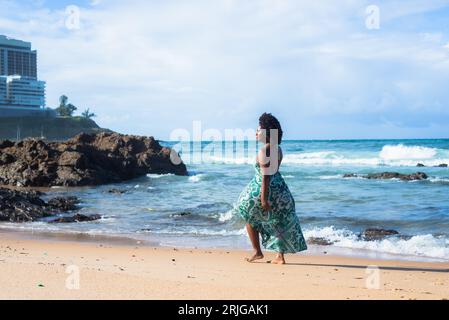 Porträt einer schönen Frau mit schwarzem, kräftigem Haar in hellgrünem, langem Kleid, die auf dem Strand spaziert. Im Hintergrund liegen die Wolken auf Felsen und das Meer. Sal Stockfoto