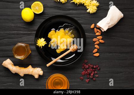 Kristallisierter Honig in einem schwarzen Teller, Honig in Gläsern, Mandeln, Zitrone, Ingwer, getrocknete Beeren auf dunklem texturellem Hintergrund, Draufsicht. Stockfoto