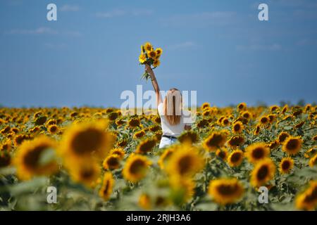 Das Mädchen steht auf einem Feld mit Sonnenblumen und hebt ihre Hand mit einem Blumenstrauß. Stockfoto