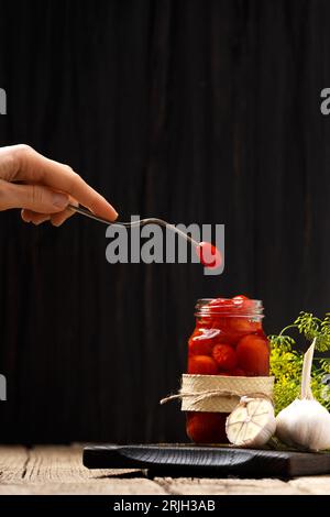 Eingelegte Kirschtomaten in einem offenen Glas auf einem dunklen Holzbrett, eine Tomate auf einer Vintage-Gabel in der Hand, dunkler Hintergrund, Nahaufnahme. Stockfoto