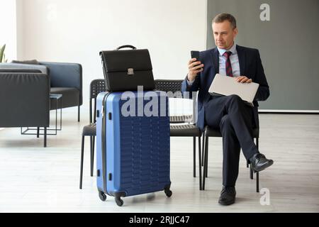Reifer Geschäftsmann mit Laptop mit Mobiltelefon in der Halle des Flughafens Stockfoto