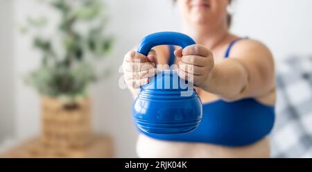 Plus Größe Frau, die Übungen mit Kurzhanteln macht, ihren Körper im Wohnzimmer stärkt, selektive Fokussierung. Stockfoto