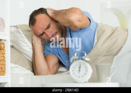 Mann, der seine Ohren bedeckt, neben dem Wecker auf dem Bett Stockfoto