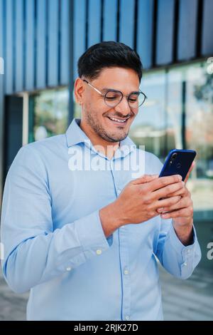 Vertikales Porträt eines hispanischen jungen Unternehmers oder Geschäftsmannes, der lächelt und eine Handy-App verwendet. Hübscher lateinischer professioneller Executive-Mann, der im Internet mit Smartphone am Arbeitsplatz surft. Hochwertige Fotos Stockfoto