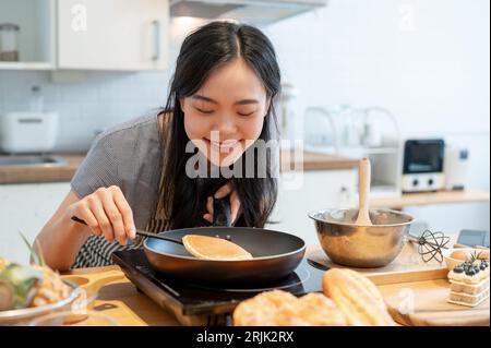 Eine glückliche und liebliche junge asiatische Frau, die einen Pfannkuchen in einer Pfanne mit einem Spatel umschlägt, kocht gerne Pfannkuchen in der Küche und bereitet ein hausgemachtes Frühstück zu Stockfoto