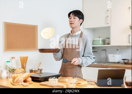 A joyful and handsome young Asian man in an apron, tossing pancakes on a frying pan, enjoys cooking pancakes in his kitchen. Stock Photo