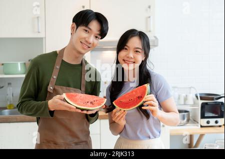Ein nettes und glückliches junges asiatisches Paar hält ein Stück Wassermelone und genießt gesundes Essen in der Küche zusammen. Stockfoto