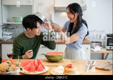 Ein hübsches und verspieltes junges asiatisches Paar tut so, als kämpft es mit Utensilien und hat Spaß beim Zubereiten von Speisen in der Küche. Lustige Kuppeln Stockfoto