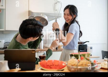 Ein hübsches und verspieltes junges asiatisches Paar tut so, als kämpft es mit Utensilien und hat Spaß beim Zubereiten von Speisen in der Küche. Lustige Kuppeln Stockfoto