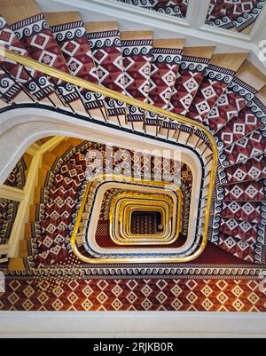 Luxuriöse Treppe in abstrakter Form mit roter Teppichabdeckung und goldenem Handlauf. Hypnotische Muster-Wendeltreppe Stockfoto