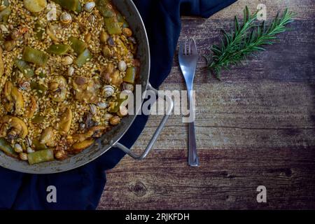 paella valenciana on cloth, with a fork and a sprig of rosemary on a wooden table, horizontal. Stock Photo