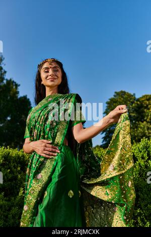 Fröhliche und moderne junge indische Frau berührt Sari mit Muster in der Nähe von grünen Pflanzen im Park draußen Stockfoto