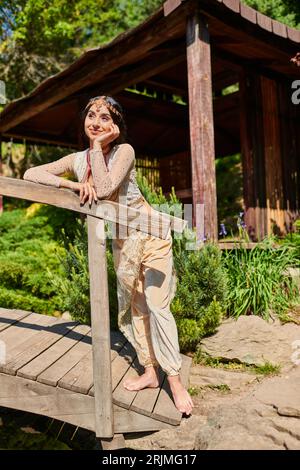 Glückliche und traumhafte indische Frau in traditioneller Kleidung, die auf einer Holzbrücke im Sommerpark wegschaut Stockfoto