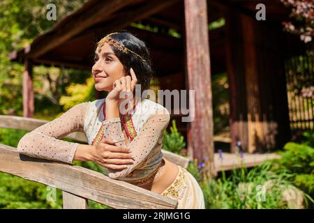 Sommerpark, traumhafte, authentische Frau im indischen Stil, die auf einer Holzbrücke lächelt und wegschaut Stockfoto