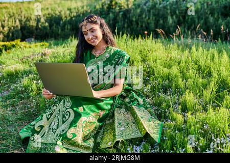 Stilvolle und lächelnde junge indische Frau in Sari mit Laptop, während sie im Sommer auf der Wiese sitzt Stockfoto