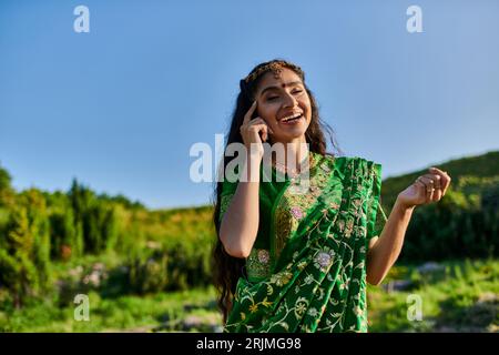 Fröhliche junge indische Frau, die das Gesicht berührt, während sie in Sari posiert, mit Landschaft im Hintergrund Stockfoto