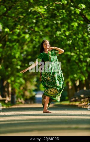 Fröhliche junge indianerin im Sari, die auf der Straße tanzt, mit grünen Bäumen im Hintergrund Stockfoto