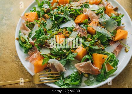 Blick von oben auf Rucolasalat mit Käse, Cantaloupe, Prosciutto, Pistazien auf weißem Teller. Goldener Hintergrund und goldene Farbgabel Stockfoto