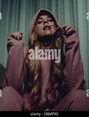 Eine junge Frau mit langen blonden Haaren und einem pinken Hoodie, der vor den weißen Vorhängen posiert Stockfoto