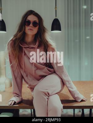 Eine junge Frau, die stilvolle rosa Loungewear und Sonnenbrille trägt und auf einem Tisch sitzt Stockfoto