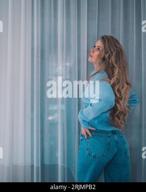 Eine junge Kaukasierin in blauem Pullover und blauer Jeans, die sich an einen Fenstervorhang lehnt Stockfoto