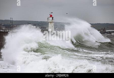Riesige Wellen stürzen in den Hafen von Newlyn ein, einige reichen bis hoch und über den Leuchtturm, der an der Hafenmauer liegt. Newlyn, Cornwall, Großbritannien Stockfoto