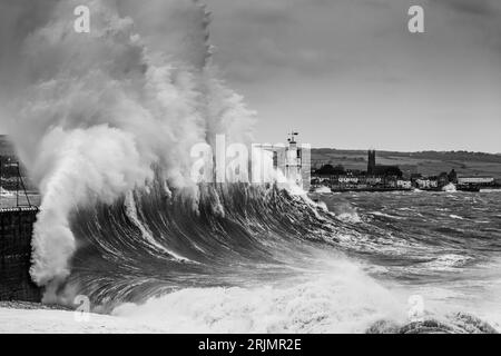 Riesige Wellen stürzen in den Hafen von Newlyn ein, einige reichen bis hoch und über den Leuchtturm, der an der Hafenmauer liegt. Newlyn, Cornwall, Großbritannien Stockfoto