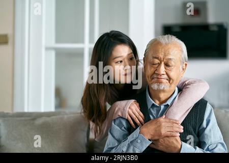 Trauriger älterer asiatischer Vater, der auf der Couch im Wohnzimmer zu Hause saß, getröstet von einer erwachsenen Tochter Stockfoto