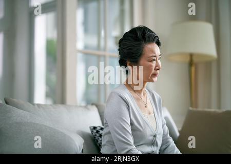 Seitenansicht einer traurigen älteren asiatischen Frau, die allein auf der Couch im Wohnzimmer zu Hause sitzt Stockfoto