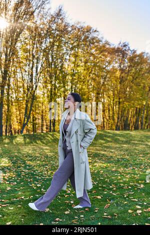 Glückliche afroamerikanische Frau im Grabenmantel, die auf Gras mit gefallenen Blättern geht, Herbst, Mode Stockfoto