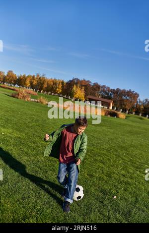 Sportlicher afroamerikanischer Junge in herbstlicher Oberbekleidung, der Fußball auf dem grünen Feld, Fußball und Herbst spielt Stockfoto