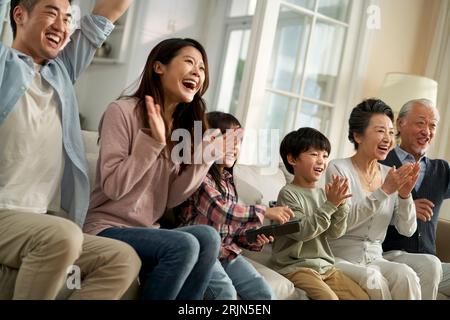 Mehrgenerationenübergreifende asiatische Familie, die im Wohnzimmer auf der Couch sitzt und sich die Fernsehserie glücklich und lächelnd ansieht Stockfoto