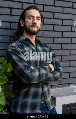 Ein hispanischer Mann, der mit überkreuzten Armen vor einem Backsteinhaus steht Stockfoto