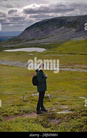 Aktive Wanderin, die an einem bewölkten Tag in schwedischer Natur den Blick über das grüne Tal, den See und den Nipfjället-Berg in Idre genießt Stockfoto