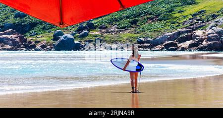 Eine junge Frau schlendert entlang der Küste und trägt ein Surfbrett unter einem großen, leuchtend roten Schirm Stockfoto