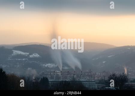 Rauch, der am Abend aus den Kaminen der Heizungsanlage kommt, Ponir-Berg mit sonnendurchfluteter Spitze und strahlendem Himmel im Hintergrund Stockfoto