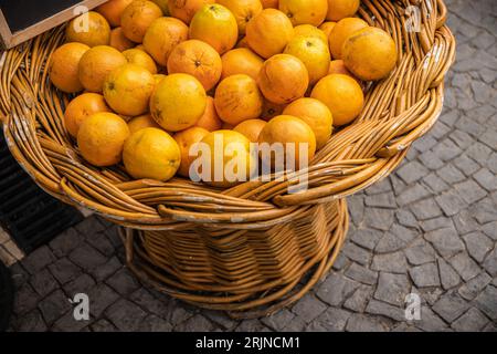 Eine Nahaufnahme eines Korbs aus Korbgeflecht, gefüllt mit Reifen Orangen, die auf einem Pflastersteinpflaster ruhen Stockfoto