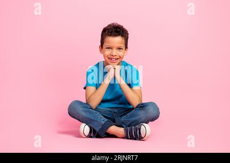 Foto in voller Länge von traumhaft süßen kleinen Jungen gekleidetes blaues T-Shirt sitzende Hände Kinn isolierter rosa Farbhintergrund Stockfoto