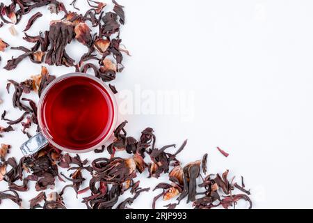 Eine bis zum Rand gefüllte Glasschale mit einem leuchtend roten Hibiskus-Tee, mit ein paar trockenen Teeblättern, die um die Tasse verstreut sind Stockfoto