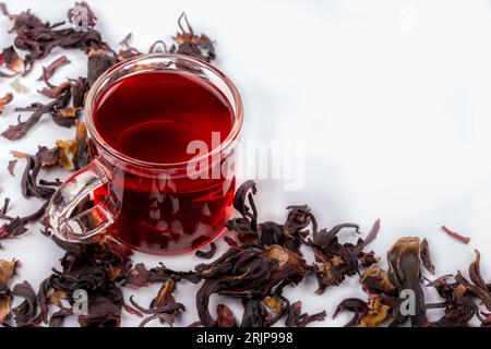 Eine bis zum Rand gefüllte Glasschale mit einem leuchtend roten Hibiskus-Tee, mit ein paar trockenen Teeblättern, die um die Tasse verstreut sind Stockfoto