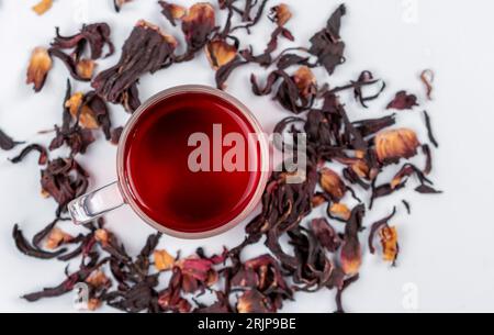 Eine bis zum Rand gefüllte Glasschale mit einem leuchtend roten Hibiskus-Tee, mit ein paar trockenen Teeblättern, die um die Tasse verstreut sind Stockfoto