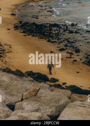 Ein Mann, der alleine an einem felsigen Strand läuft und einen Wanderstock ergreift, während er in den Horizont blickt Stockfoto