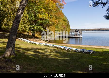 Ein malerischer Blick auf Liegestühle am Strand mit einem See im Hintergrund und umliegende Bäume an einem sonnigen Herbsttag in Muskoka, Ontario Stockfoto