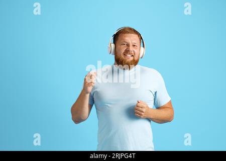 Bärtiger rothaariger Mann, der Musik über Kopfhörer vor blauem Studiohintergrund hört. Fröhliche Emotionen Stockfoto