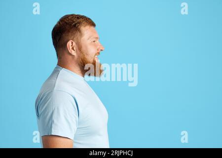 Profil, Seitenansicht eines bärtigen Mannes in einem lässigen weißen T-Shirt, das gerade vor blauem Studiohintergrund aussieht Stockfoto