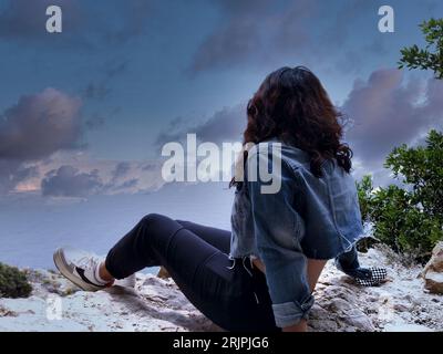 Eine junge Frau, die auf einem Felsen sitzt und in der Dämmerung zum Himmel blickt Stockfoto